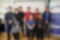Photograph shows eight adults posing at an ENOKU event or workplace gathering. Participants wear casual and formal clothing, with ENOKU banners, logo,​