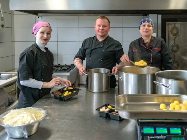 Three people standing in a commercial kitchen with pots and displaying the food they've made.​