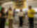 A group of six people stand attentively in a store aisle filled with organised plastic containers. One man in a yellow shirt is speaking to them.
