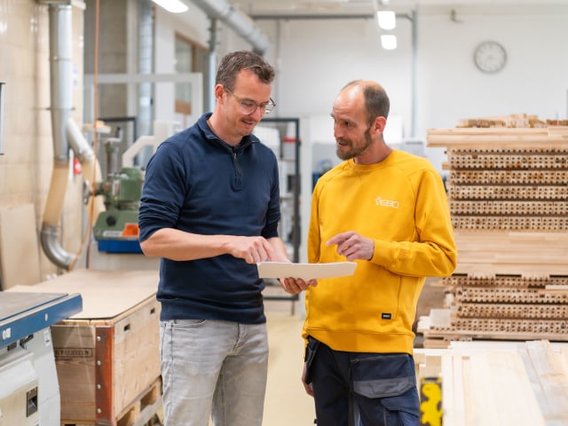 Two men standing together in a factory planning together. One is wearing blue, the other a yellow VEBO branded jacket. Wooden pallets are in the background.